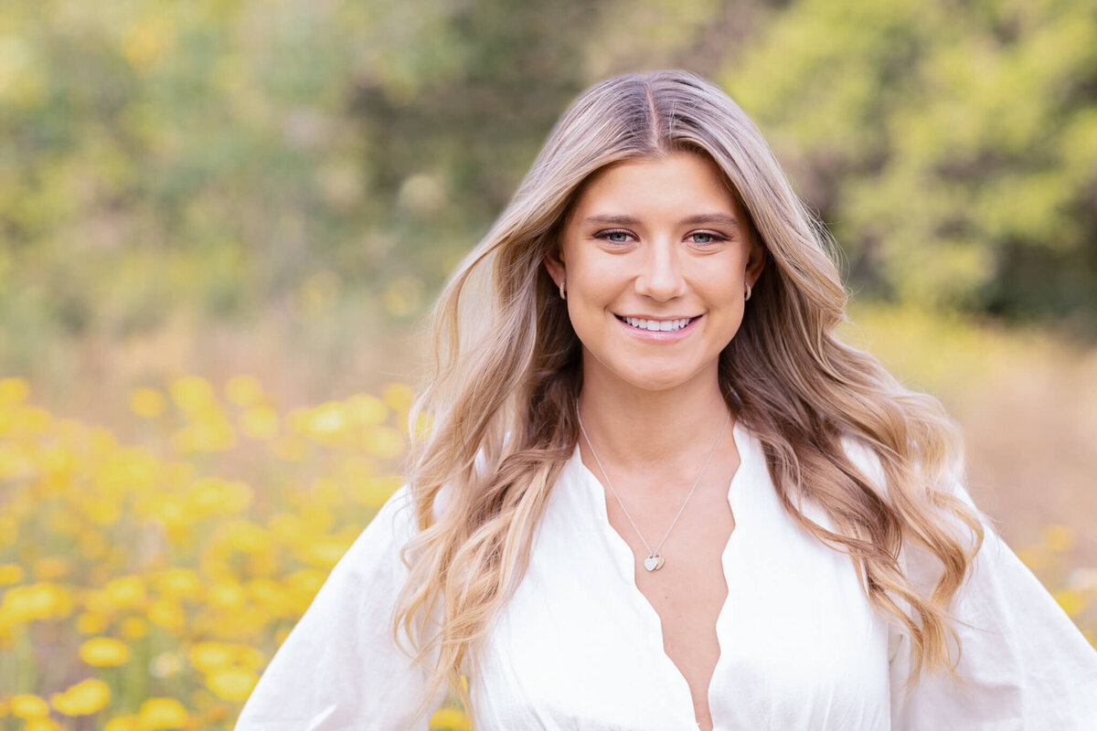 A woman with long blonde hair is smiling and standing outdoors in front of a blurred background of yellow flowers and greenery. She is wearing a white blouse and a necklace.