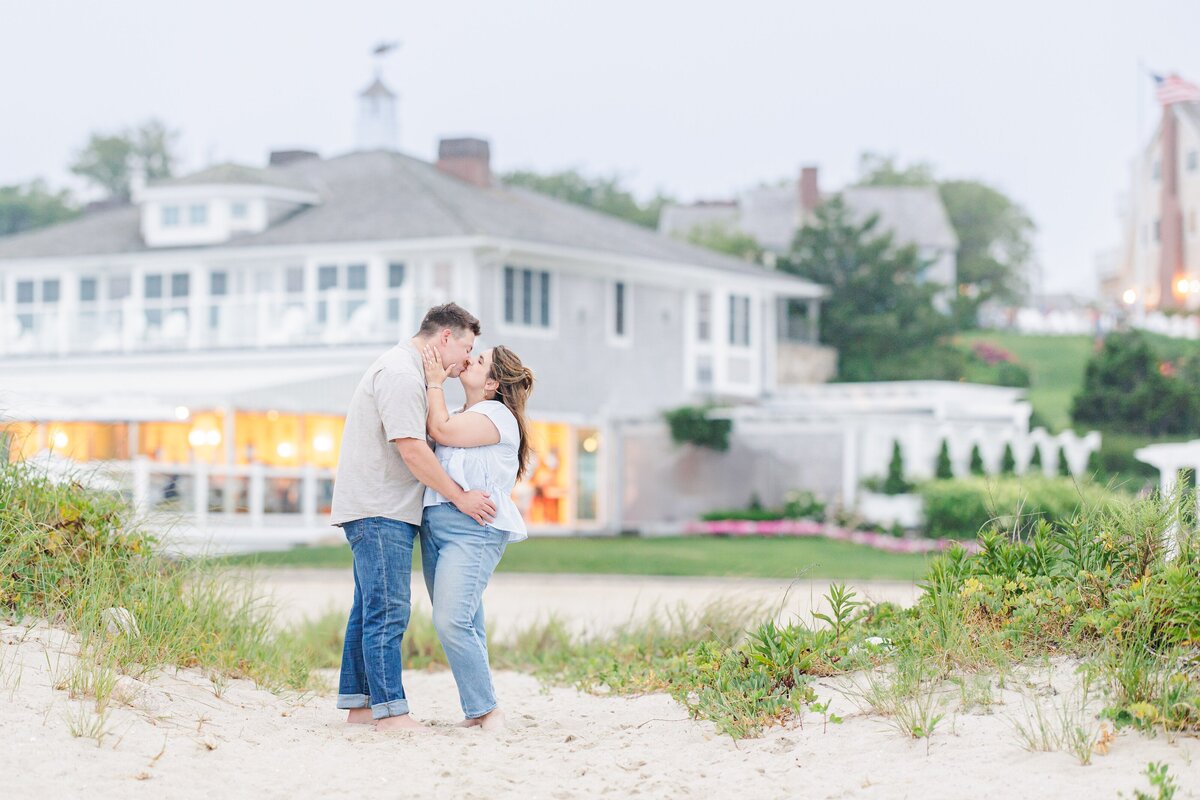 Couple kissing on the sandy beach with tall grass in the foreground and the warm lights of Chatham Bars Inn glowing behind them in Chatham, Massachusetts — representing the portfolio of a Cape Cod engagement photographer.