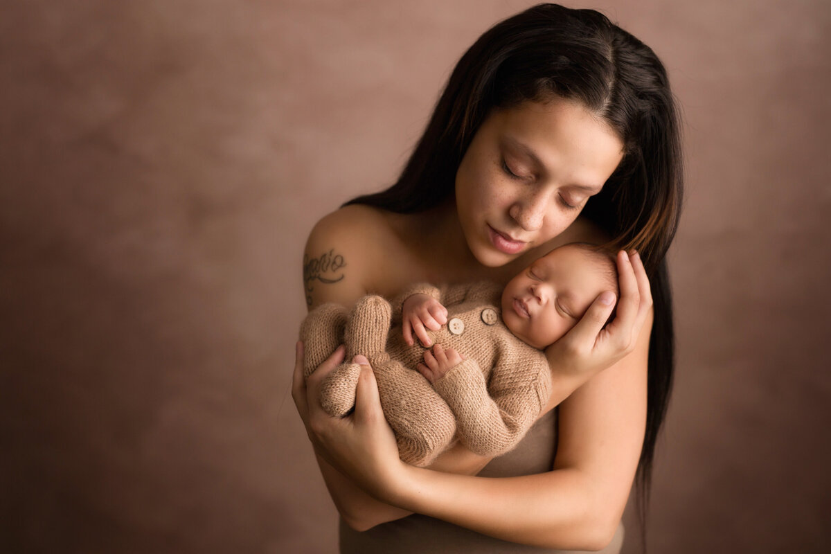 African american mom holds baby in her arms.  Mom is looking down and softly lit on a taupe background.  The baby boy is wearing a tan knit romper.  Mom has her eyes closed and is cradling baby's head in her hand.  Her other hand is holding his feet.