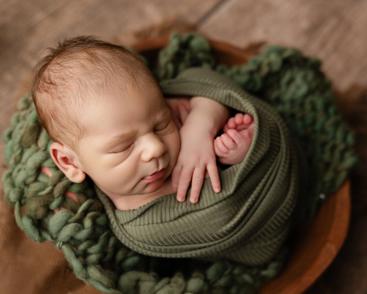 Close-up of a newborn baby boy wrapped in forest green knits, resting in a wooden bowl with textured layers. Captured by Inland Empire newborn photographer Kaitlyn Dawn Photography.