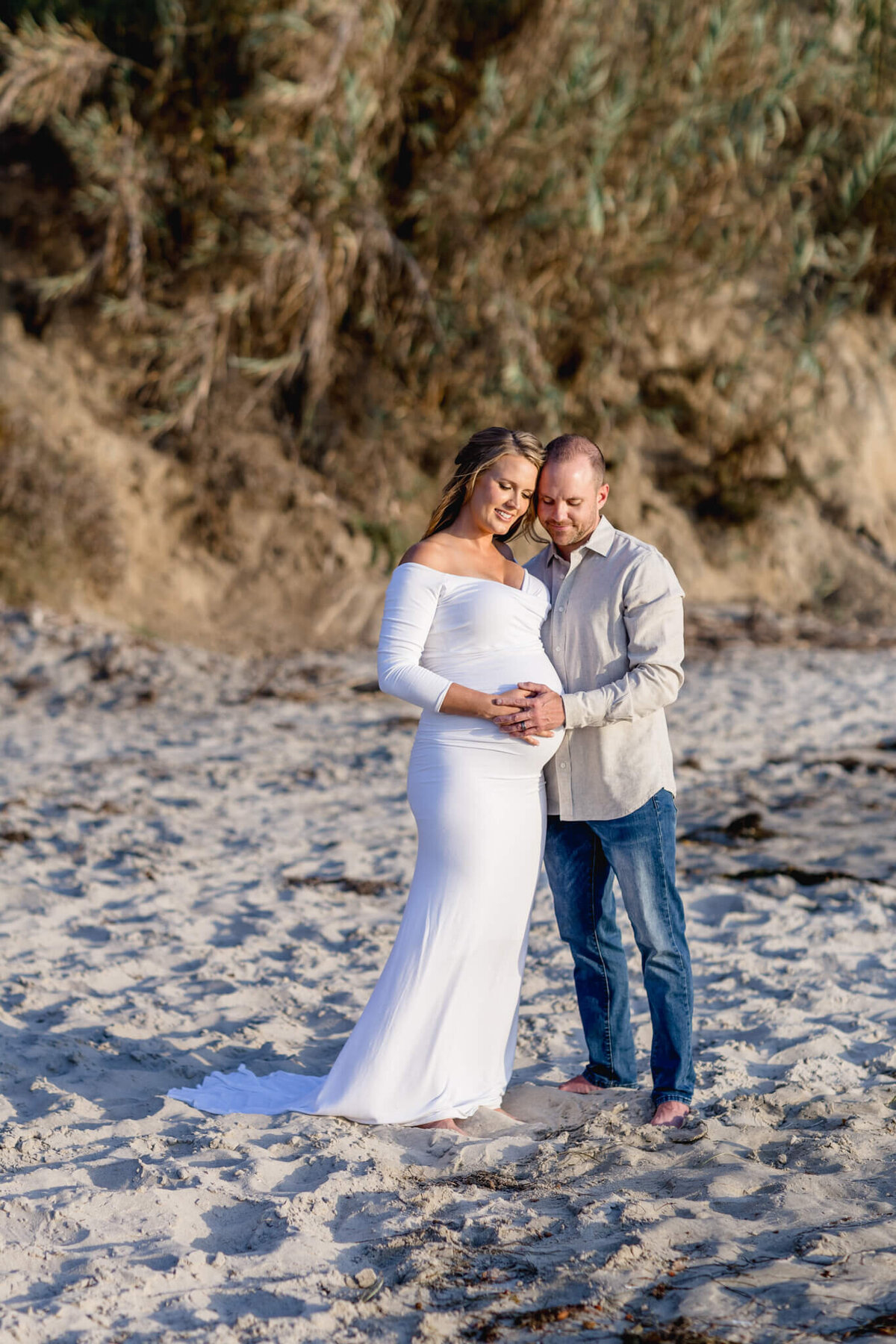 A couple stands on a sandy beach, surrounded by trees. The woman in a white dress cradles her baby bump, while the man in a beige shirt and jeans embraces her, both looking content.