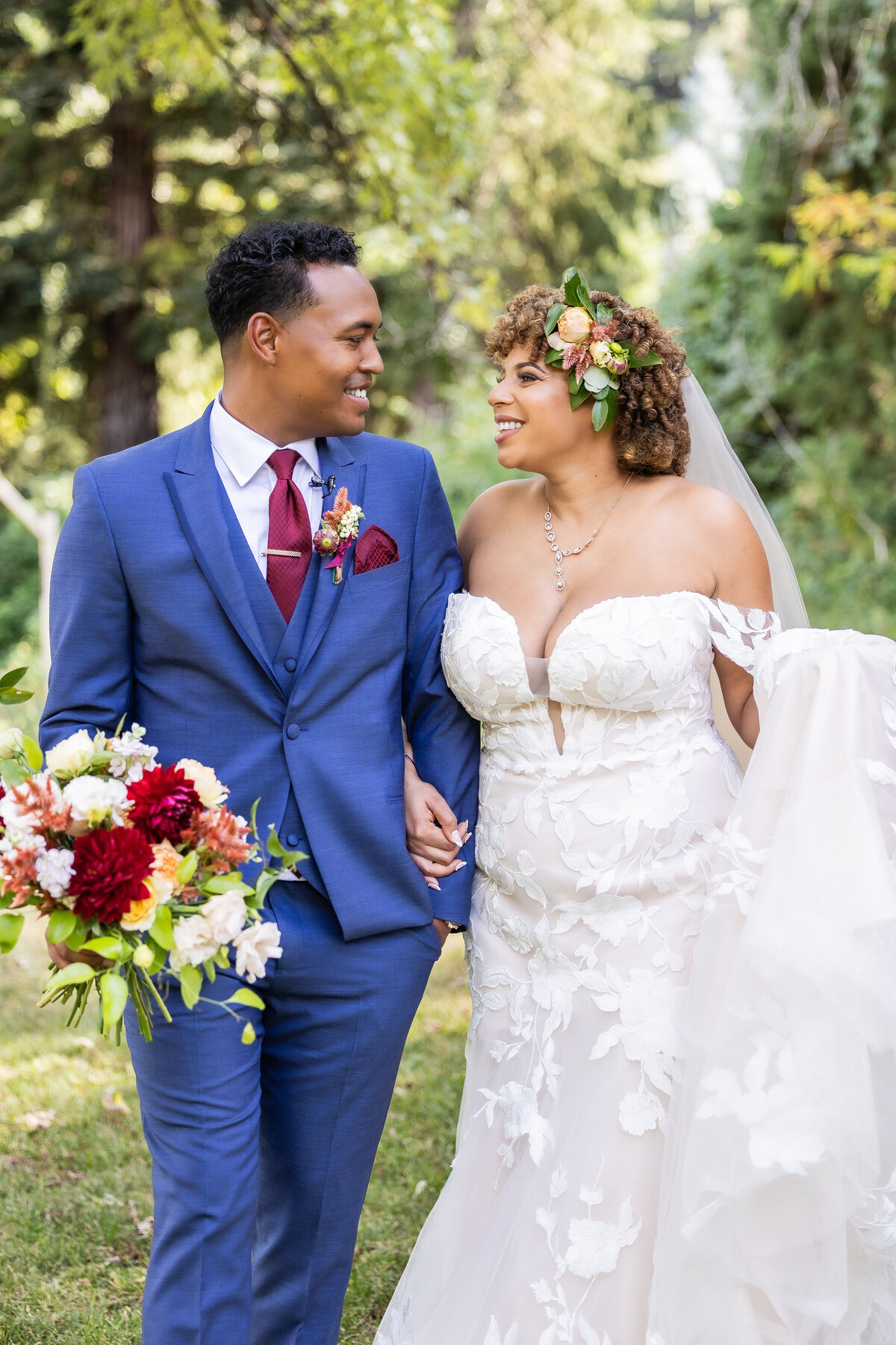 Bride and groom walk while looking at each other. Groom holds the bride's bouquet and bride holds the bottom of her dress and they are surrounded by greenery in the background. Photo by wedding photographer sacramento, ca philippe studio pro.