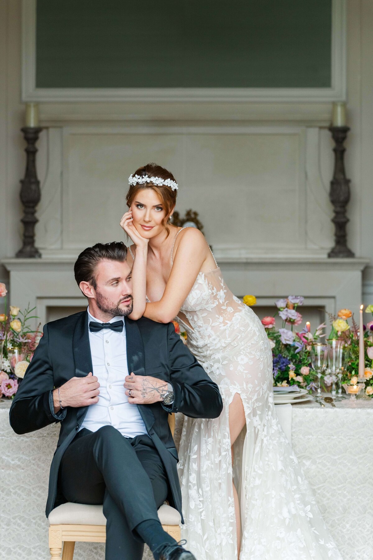 bride and groom posig, she is looking at the camera while standig, he is sitting down lokking to the side