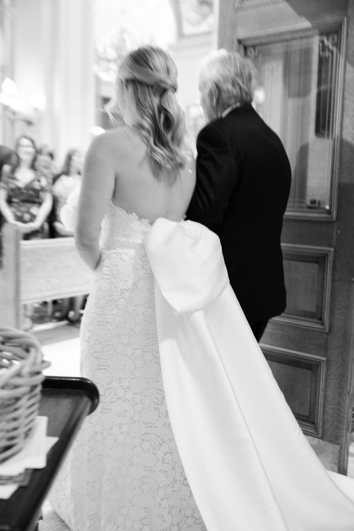 A black and white photo captures the bride, wearing a strapless lace gown with a statement bow, walking arm-in-arm with her father toward the altar. 