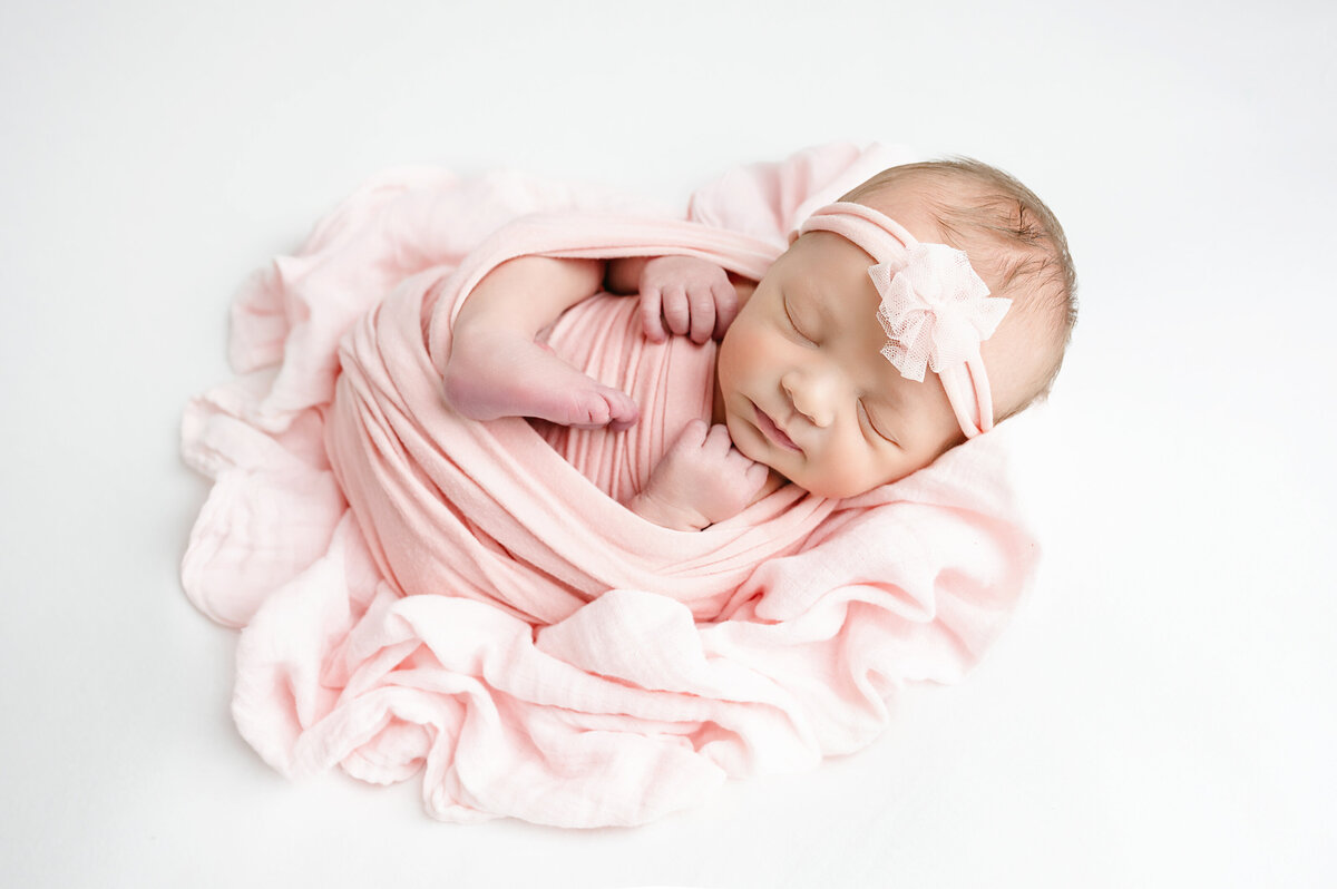This adorable newborn baby girl photography session in Pittsburgh, PA features a peaceful baby wrapped in a soft pink blanket with a matching headband. The gentle colors and serene expression make this a perfect newborn portrait.