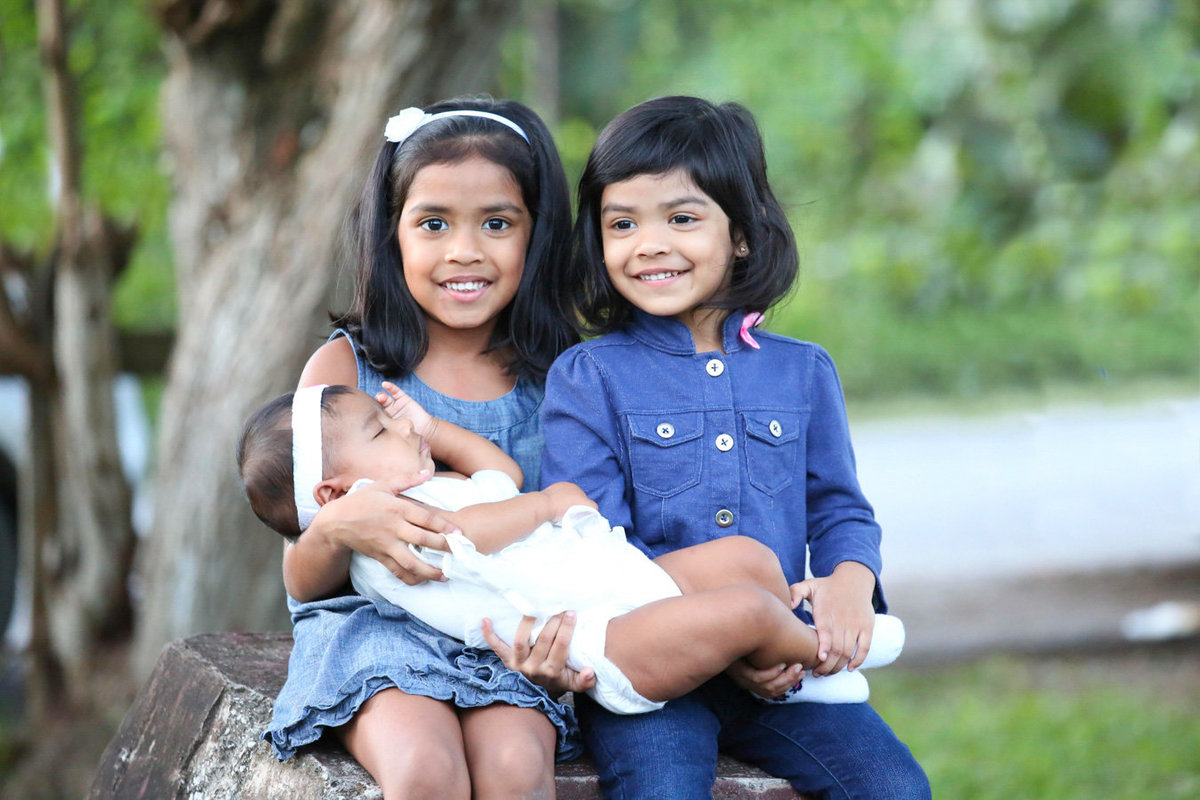 Two young girls dressed in blue holding a baby in white. Photo by Ross Photography, Trinidad, W.I..