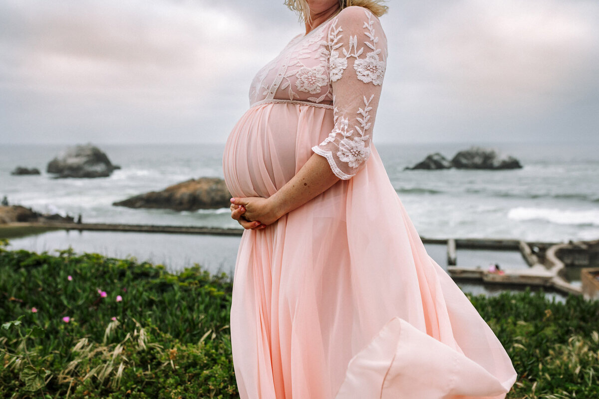 pregnant woman in flowing pink dress blowing in the wind at san francisco beach with pacific ocean in the background