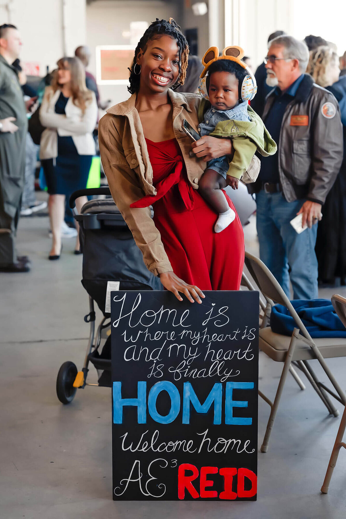 A woman holds her infant son and a welcome home sign while waiting for her husband to return from deployment.
