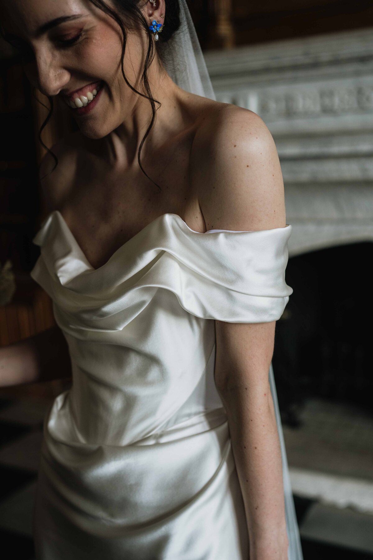 A bride is photographed wearing a satin off-the-shoulder Vivienne Westwood wedding dress. The dress has a draped neckline and she smiles warmly as she’s photographed by her wedding photographer. Her dark hair is styled in loose curls, complemented by pearl earrings. The room features a marble fireplace and checkered floor tiles.