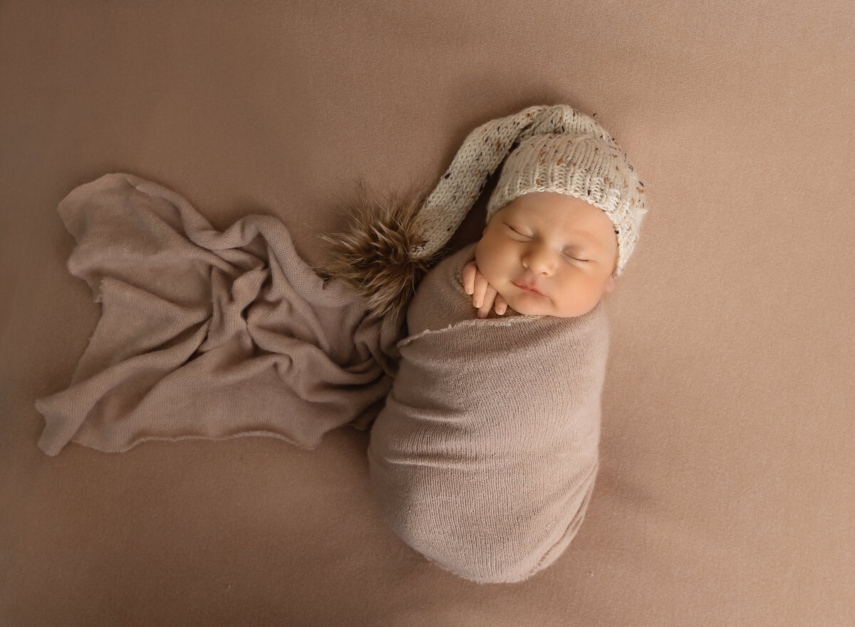 baby boy wrapped in taupe for his brooklyn newborn photoshoot. Baby is wearing a knit cap that trails beside him. He is sleeping and his fingers are peeking out of the swaddle. Captured by top Brooklyn newborn photographer Chaya Bornstein