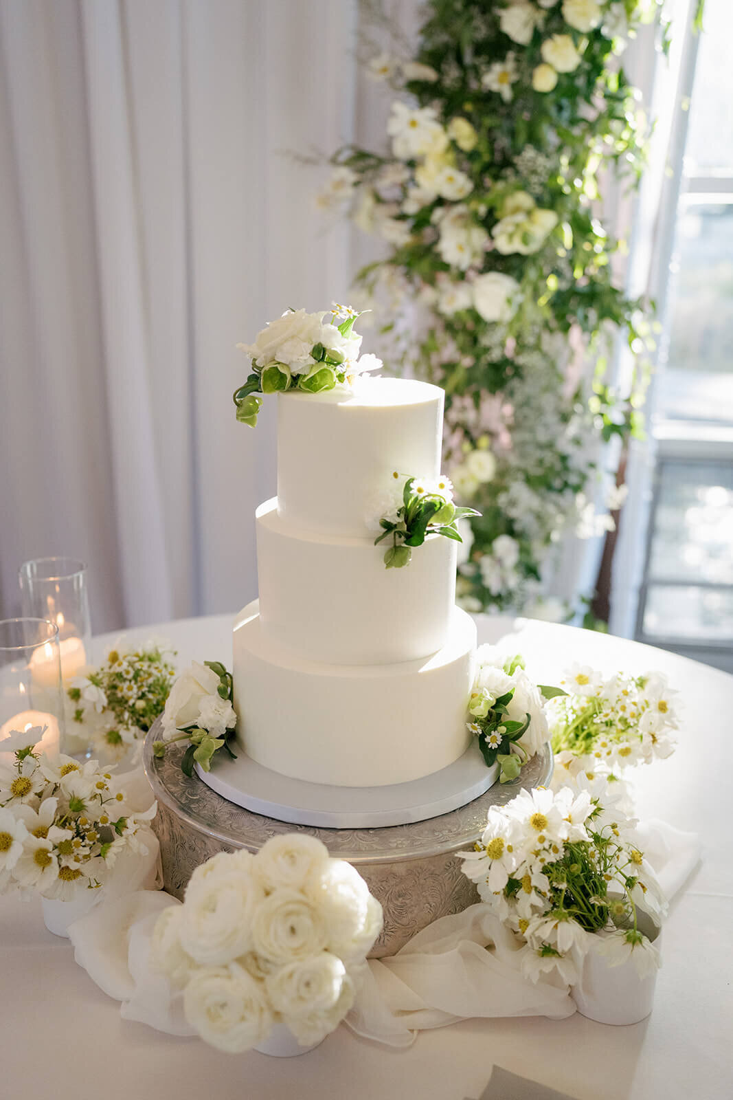 A simply classic white wedding cake with floral and greenery details atop a cake table in a wedding reception venue filled with natural light at Aspen Meadows Resort.