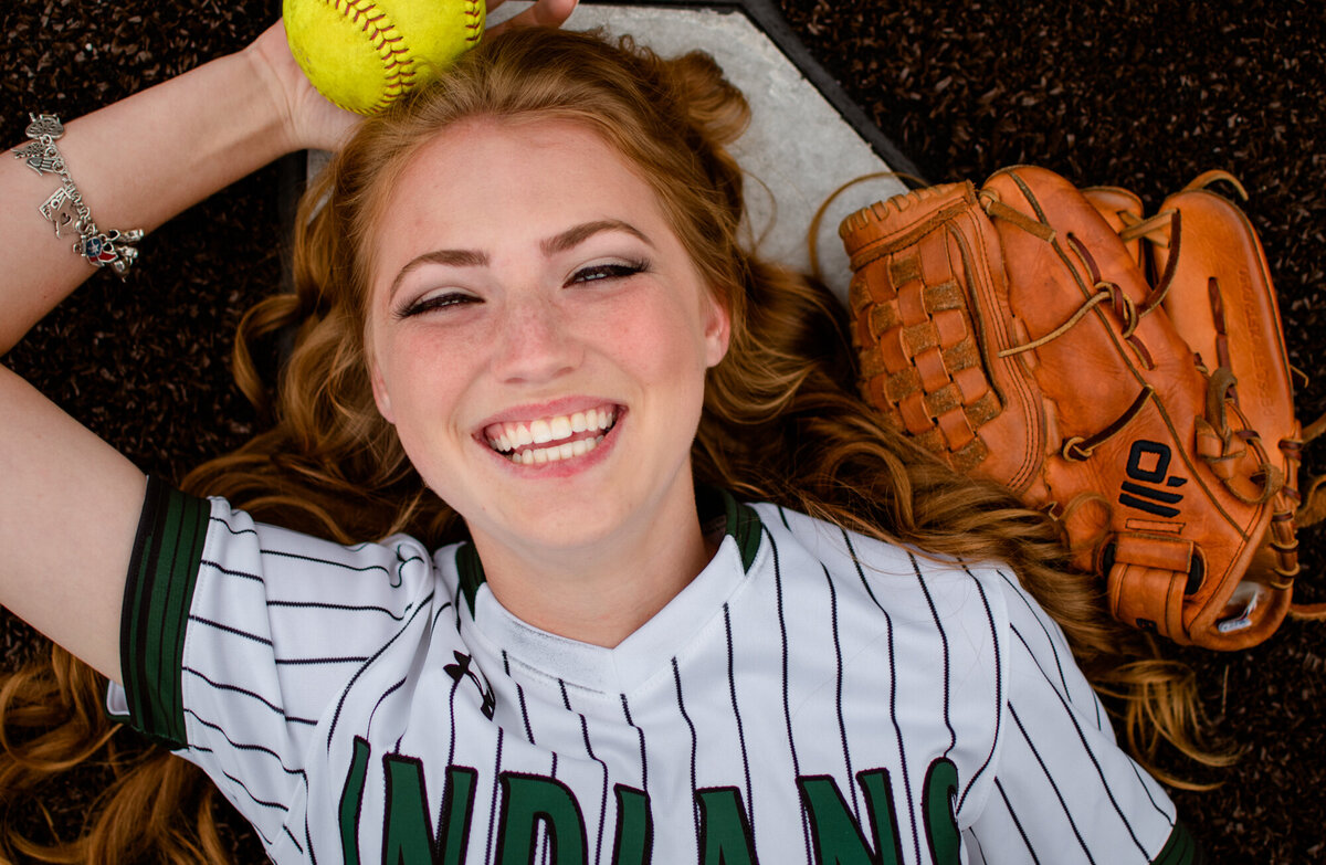 A high school senior is laying on a softball field wearing her uniform and holding a ball and her glove.