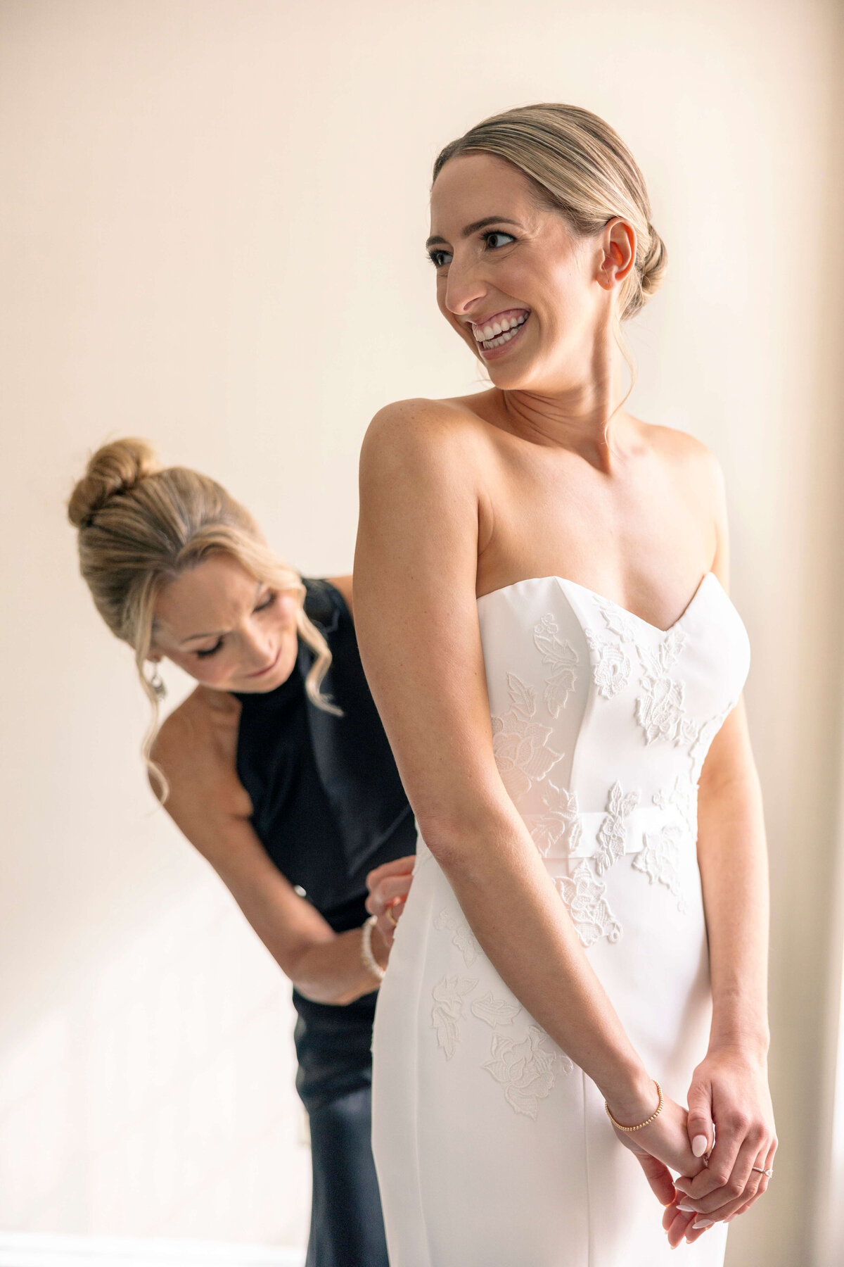 A bride wearing a strapless white gown smiles as a woman in a black dress helps adjust her gown. The background is a softly lit, neutral-colored room.