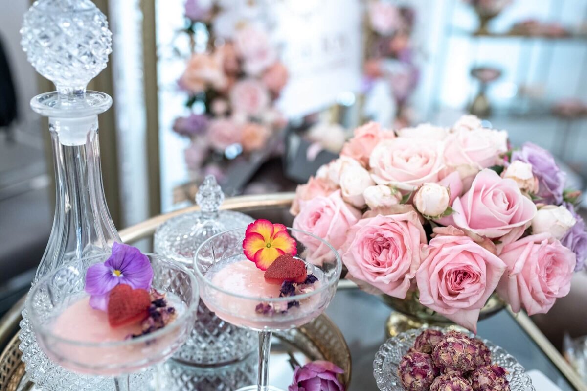 Floral cocktails in crystal glasses with a crystal decanter on a glass table with pink roses and rose bon bon desserts