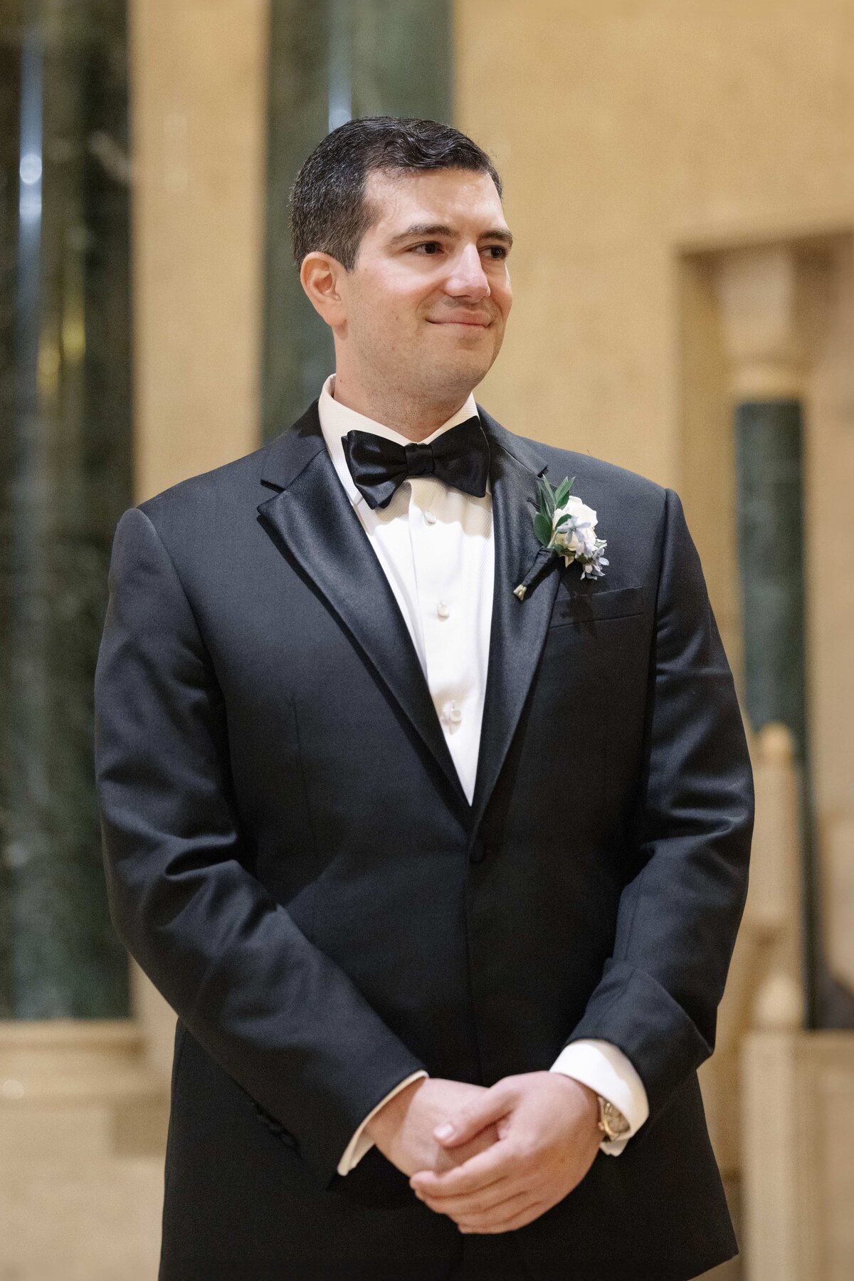 A man in a black tuxedo with a bow tie and floral boutonniere stands smiling indoors. He has short dark hair and is positioned against a background of marble-like columns.