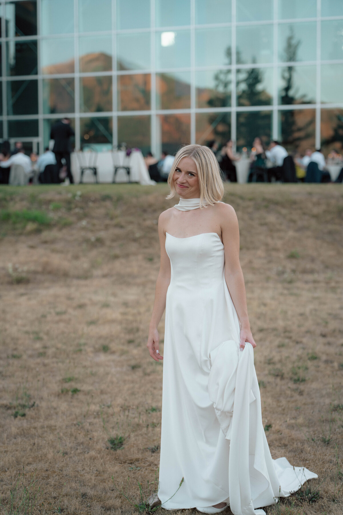 Bride in strapless gown posing on grass outside glass venue at sunset