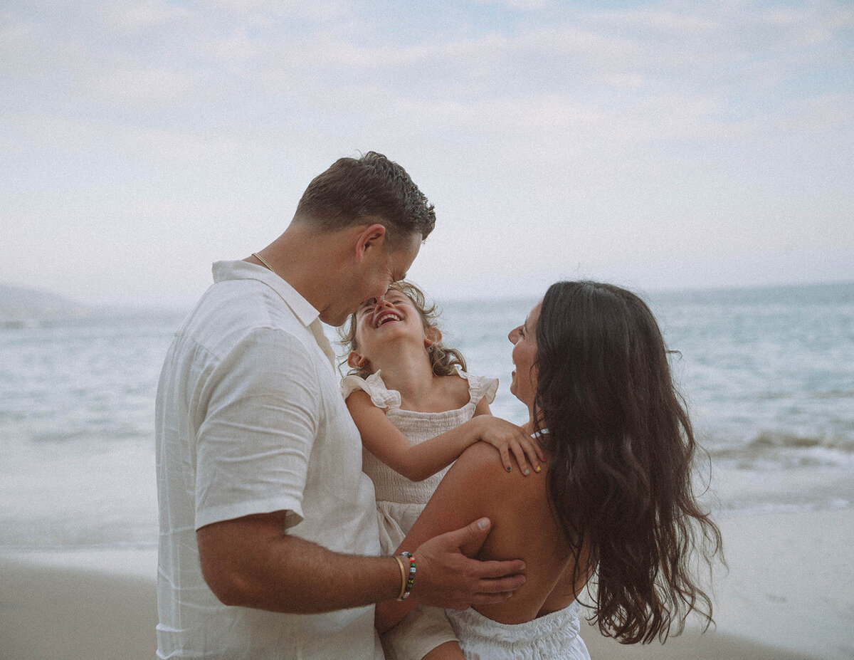 Father and mother holding their daughter at Laguna Beach, laughing together during a candid family photo session.