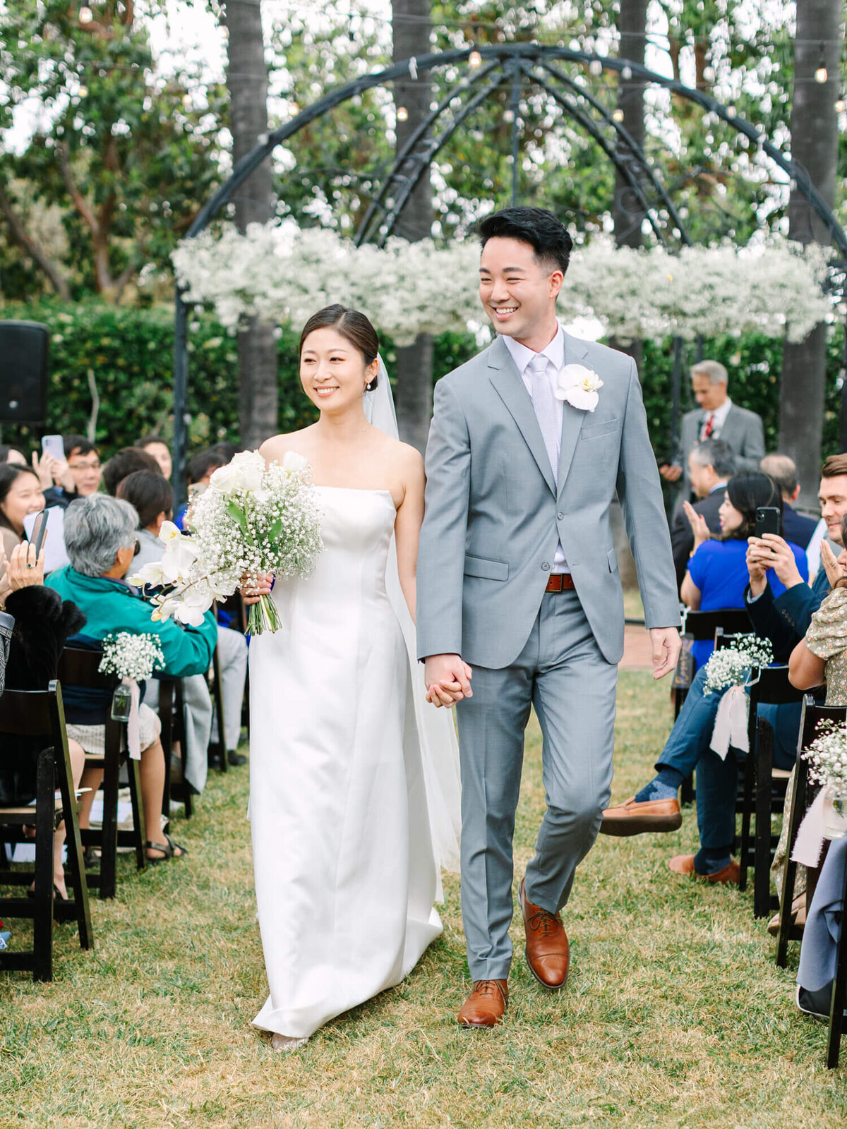 Bride in a white gown and groom in a light gray suit, smiling and walking hand in hand down an outdoor aisle amid cheering guests. Romantic and joyful.