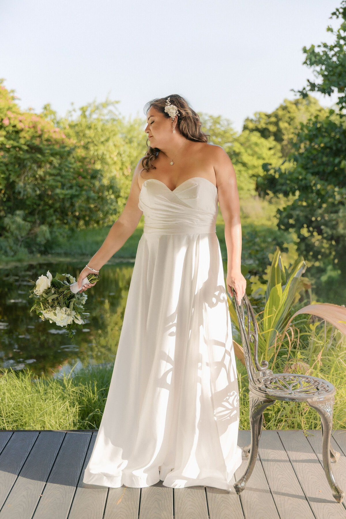 Bride in a strapless white gown holding a bouquet and smiling under golden sunlight at a lush garden venue in Miami, Florida.