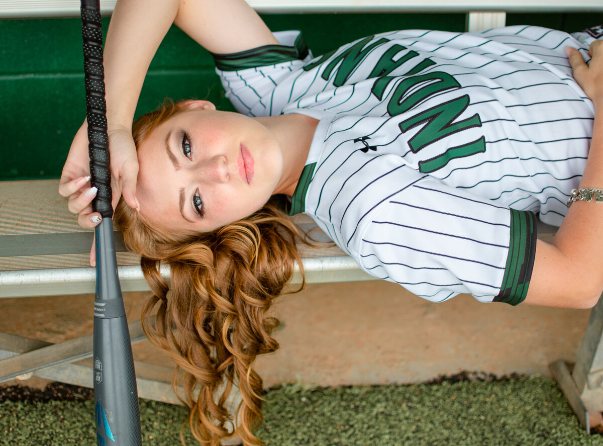 A Santa Fe High School softball player lays on a bench in a dugout holding her bat.