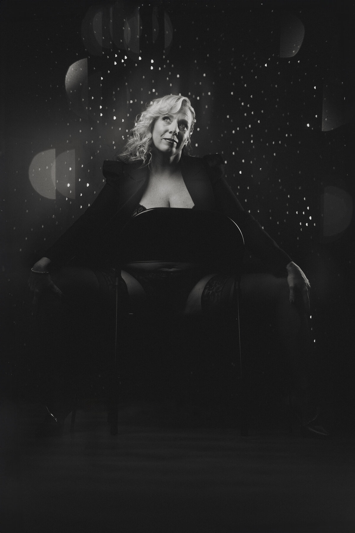 Glamorous seated pose in a black blazer and lace lingerie, celebrating joy and confidence with editorial-style lighting.