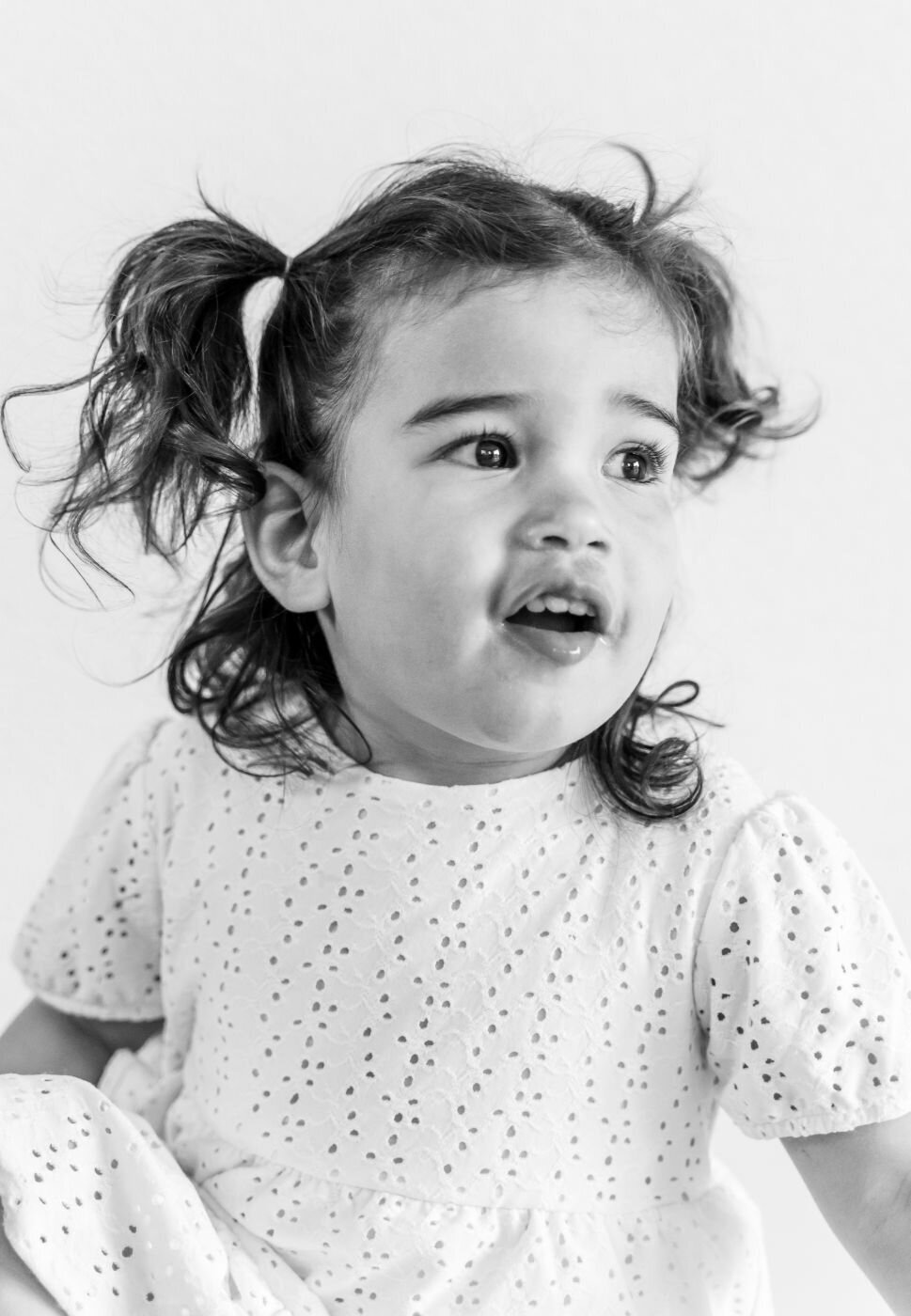 A black and white portrait of a little girl with a happy, surprised expression. She has curly brown hair styled in two pigtails. She is wearing a white dress with eyelet cutouts and is looking off to the side with her mouth slightly open. The background is a simple white.