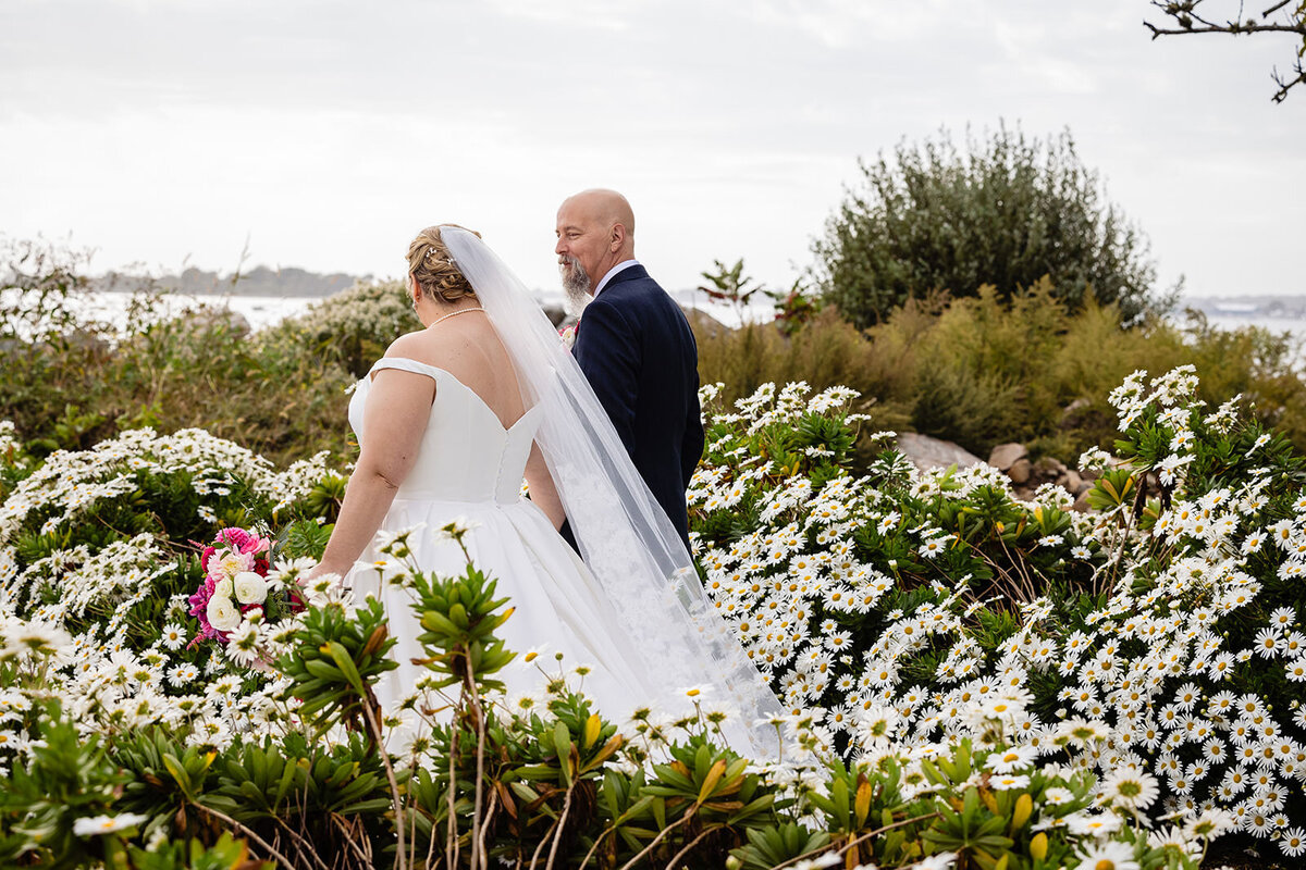 A bride and groom walking through a garden, with the bride holding her dress and a pink bouquet, and the ocean in the background