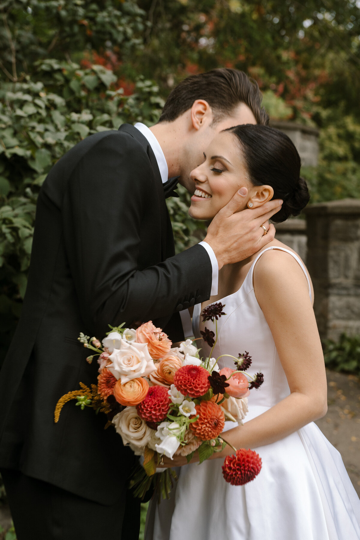 A couple embrace in the garden of Theatre St James. The bride holds bright orange flowers.