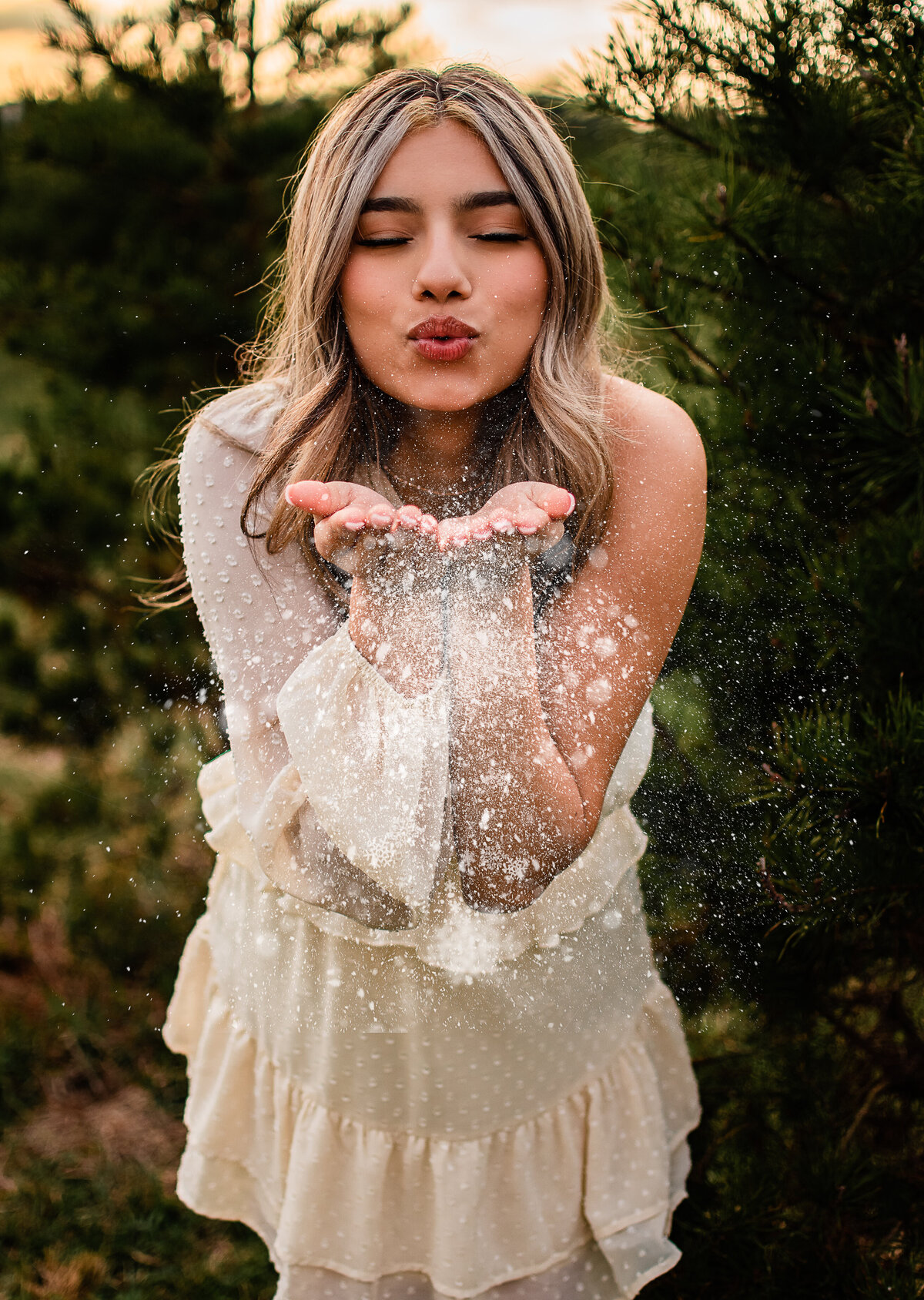 A senior wearing a white dress blows snowflakes at the camera at a tree farm.