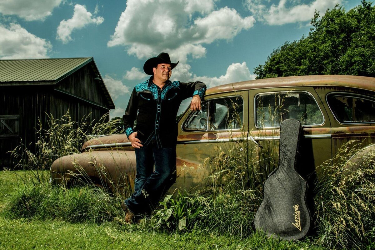 Country Music Photography Ricky Cook standing beside old car parked in overgrown grasses in front of barn Gibson guitar case leaning on car beside him