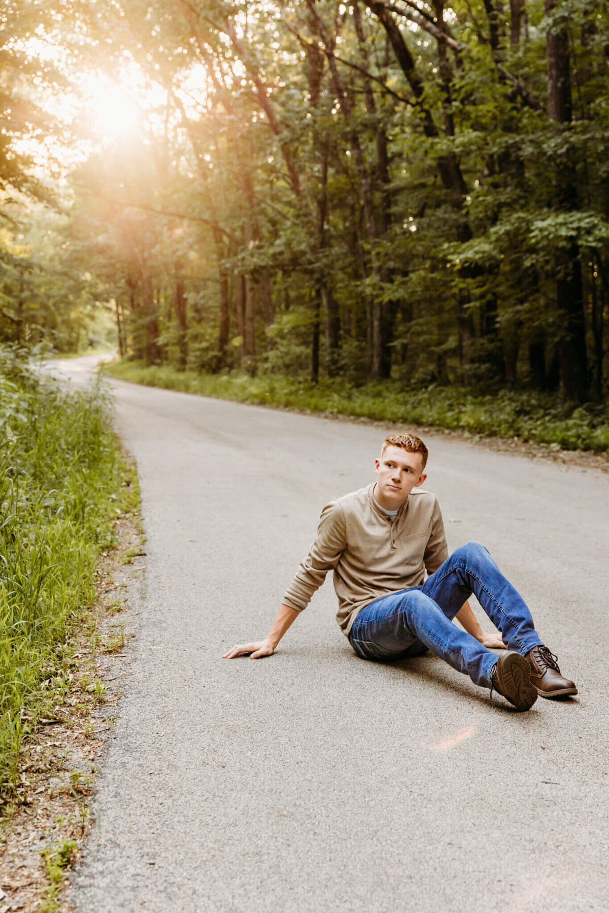 senior boy sitting in a winding road with the sun peeking through the trees