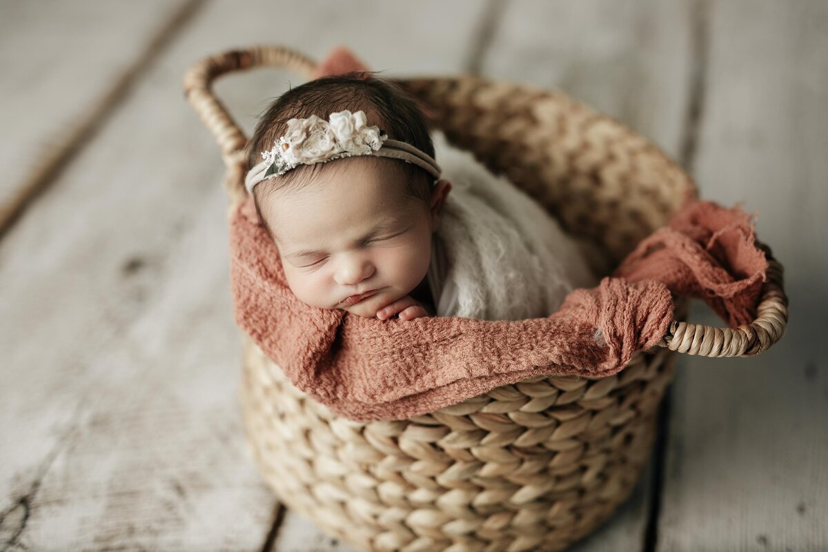 Studio newborn photography - baby sleeping in basket on an apricot blanket. Baby is in a cream wrap with a delicate floral headband. Baby's fingers are peeking out over the edge of the basket.