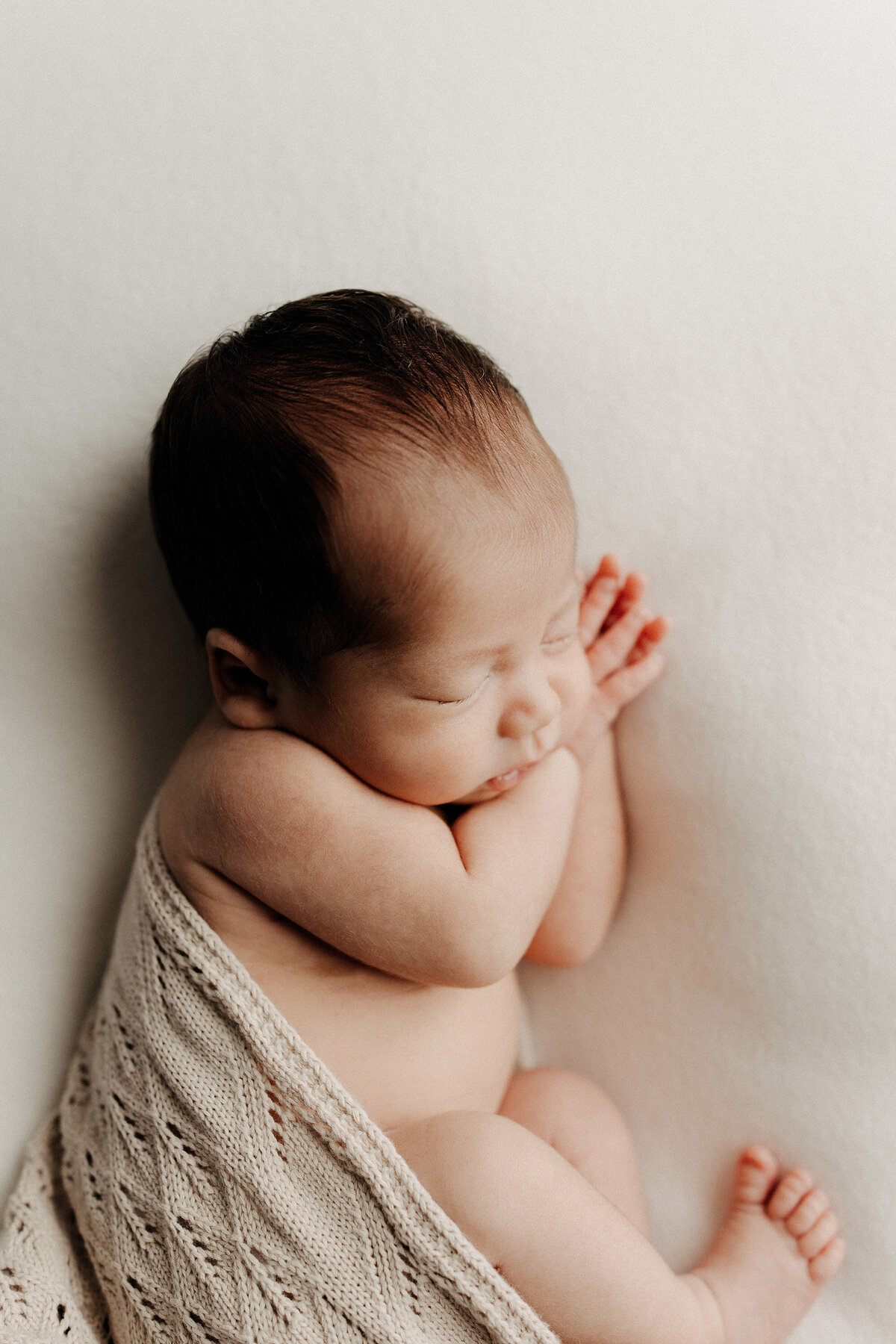 Side profile of a newborn baby sleeping curled up on a cream surface, partially wrapped in a beige knit blanket.