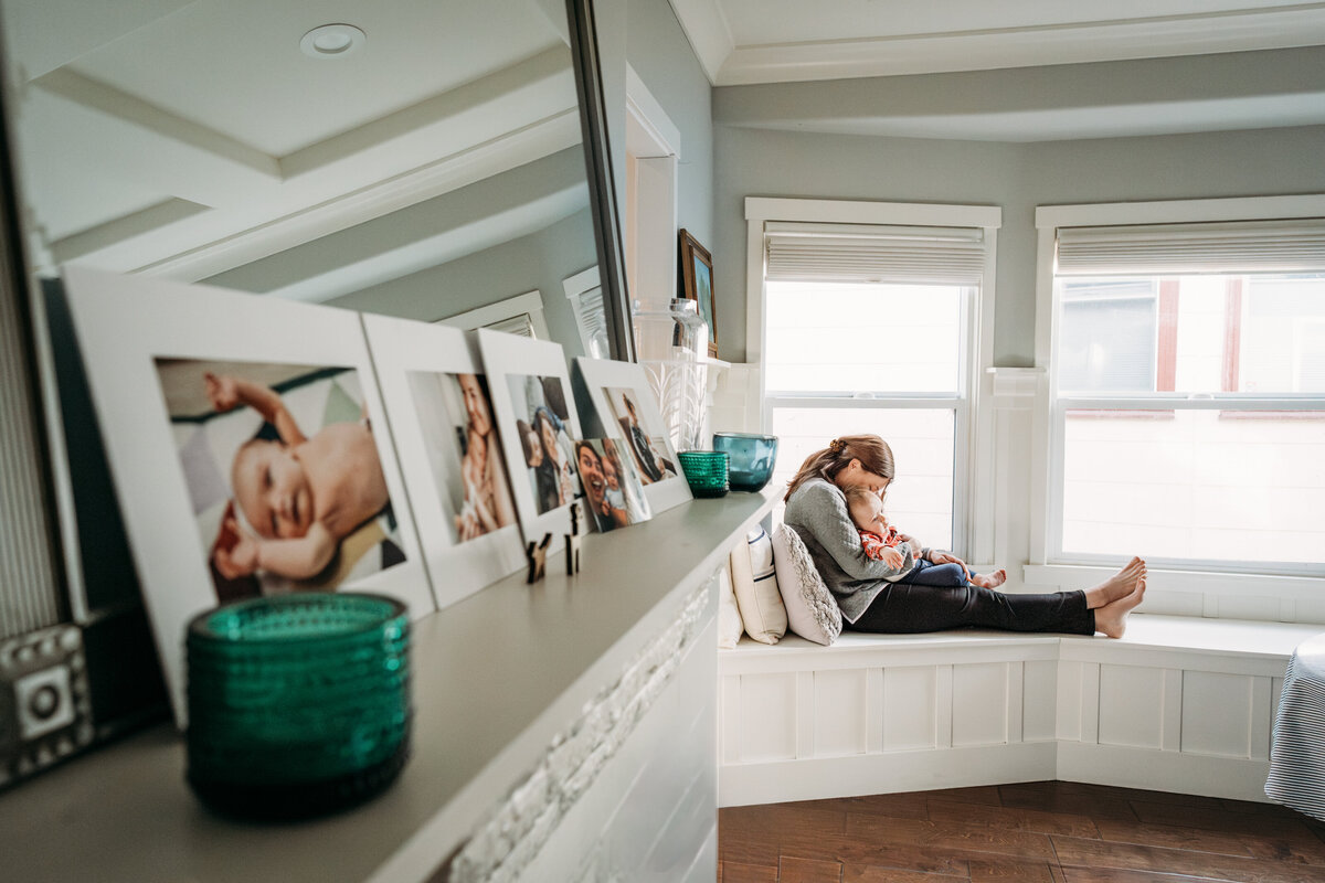 A woman sits in a bay window holding and snuggling her baby in her lap with her legs stretched out on the window bench.  the room is filled with natural light.  The perspective is from the far side of the mantle where newborn photographs are displayed.   There are dark green textured glass candle holders on the mantle.