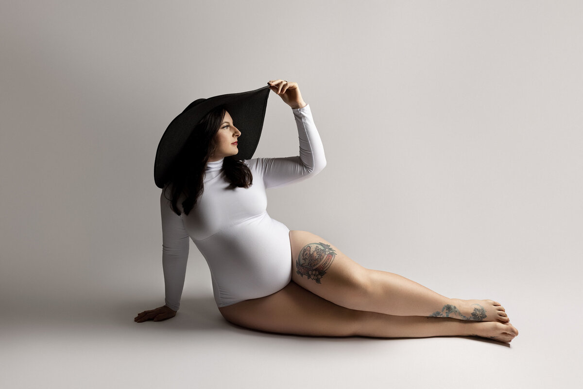 Maternity photos captured by top London, ON Maternity Photographer Amy Perrin-Ogg. Woman in white bodysuit is sitting on her side on a white backdrop wearing a wide brimmed hat. One hand is touching the top of her hat, the other is resting on the ground.