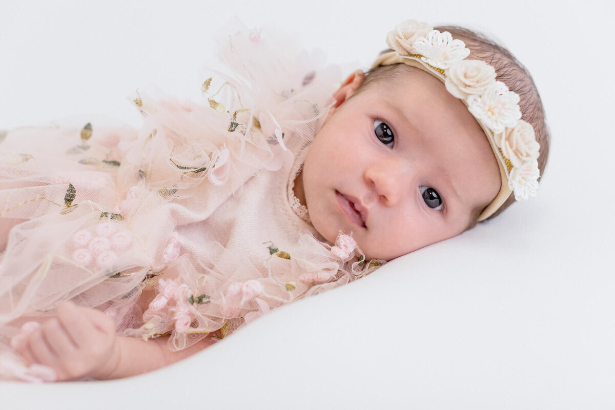 A baby wearing a floral headband and a soft pink dress with delicate floral embellishments lies on a white surface, looking directly at the camera.