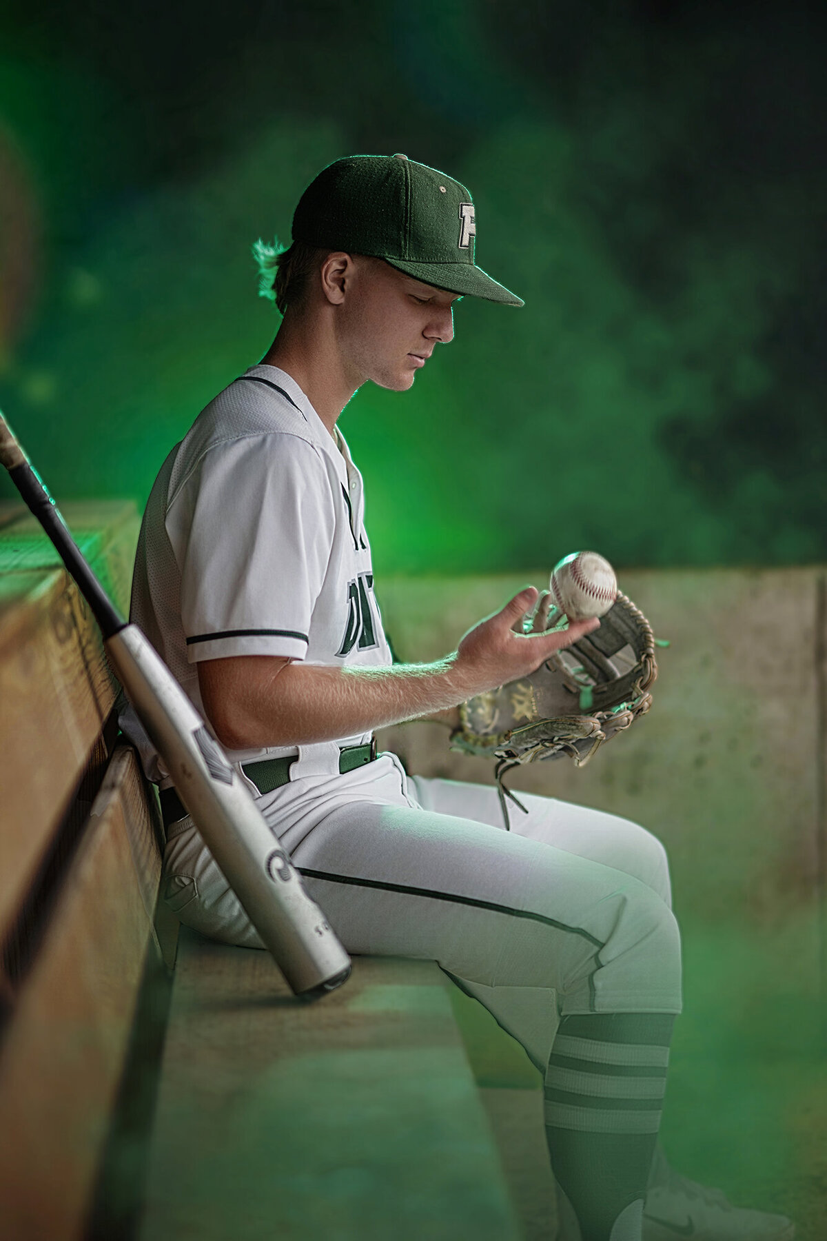 A high school baseball player sits in the dugout playing with a ball and glove with a green haze