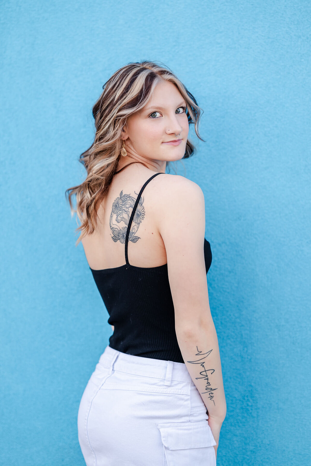 A high school senior, wearing a black top and white denim skirt, looks over her should. You can see the tattoos on her shoulder and arm. She is in front of a blue wall.