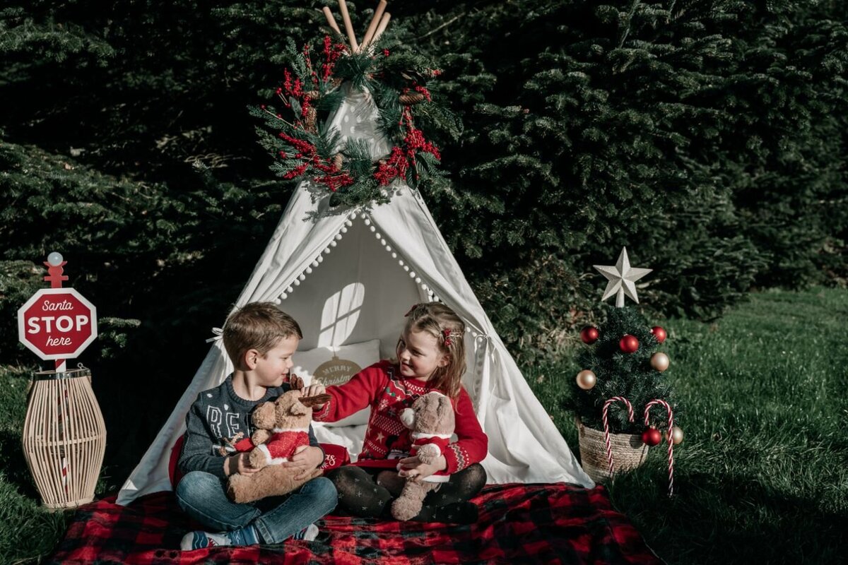 Kids in outdoor Christmas setup – Two children in festive sweaters sitting on a red-plaid blanket with teddy bears inside a small holiday teepee decorated with greenery and ornaments.