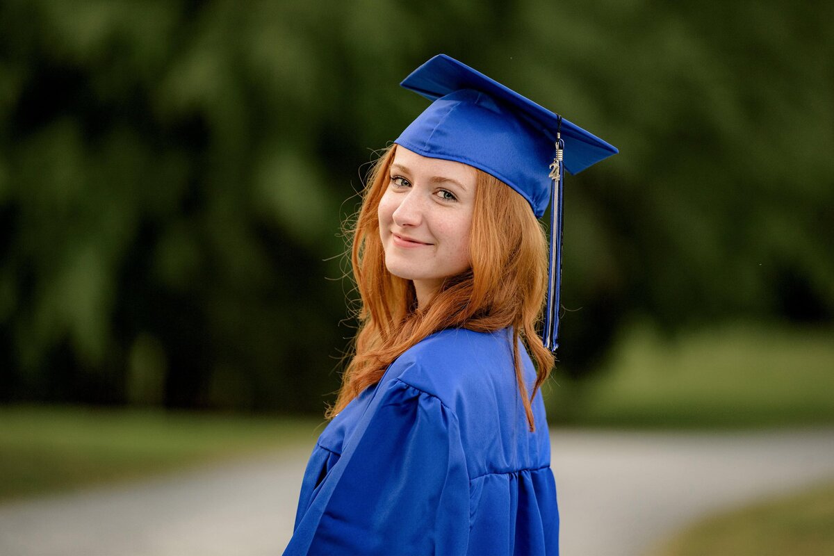 Redheaded parkwood high school graduate poses in her cap and gown at a local waxhaw, nc park while looking over her shoulder