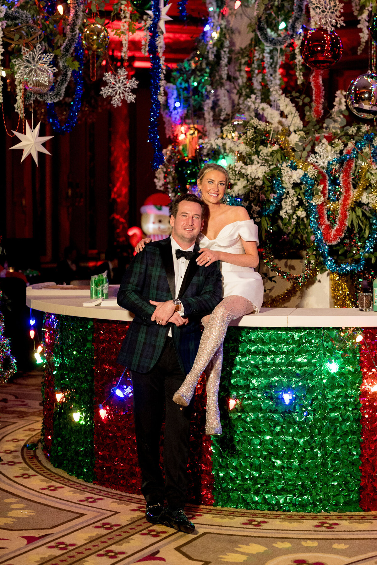 Bride in a stylish white dress and groom in a tartan suit posing with festive holiday decorations at their wedding party.