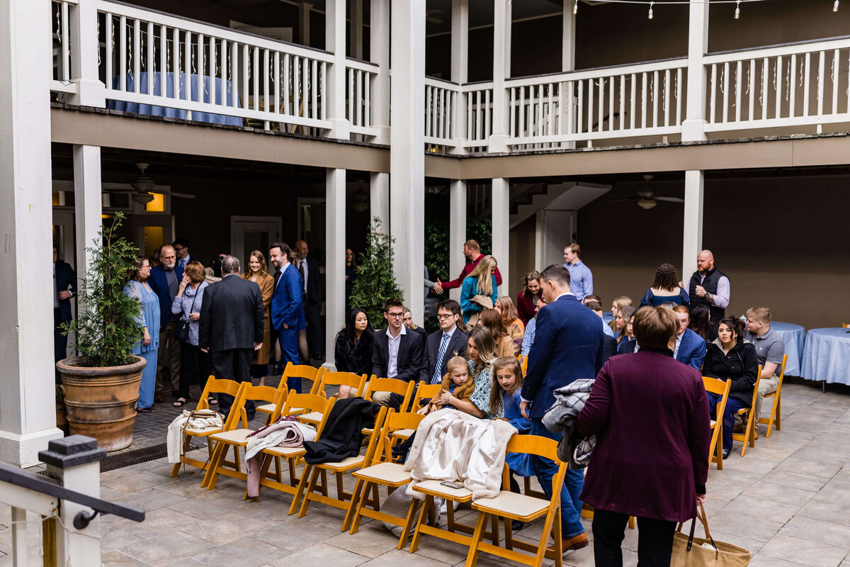 A group of people, some standing and some seated on wooden chairs, gather in a courtyard with two-story white railings, preparing for an event. The atmosphere is lively and social.
