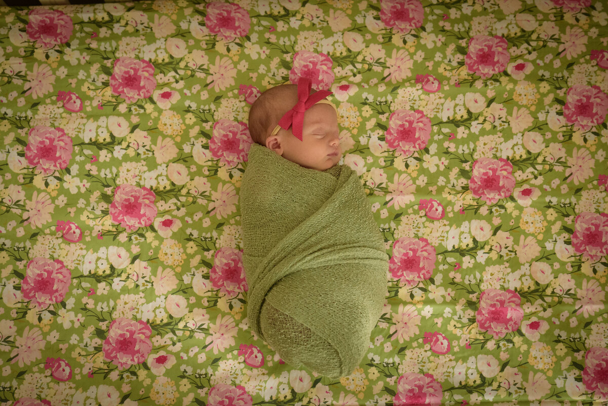 Portrait of newborn baby girl wearing a sage green wrap and pink bow in the nursery of her home in Green Bay, Wisconsin