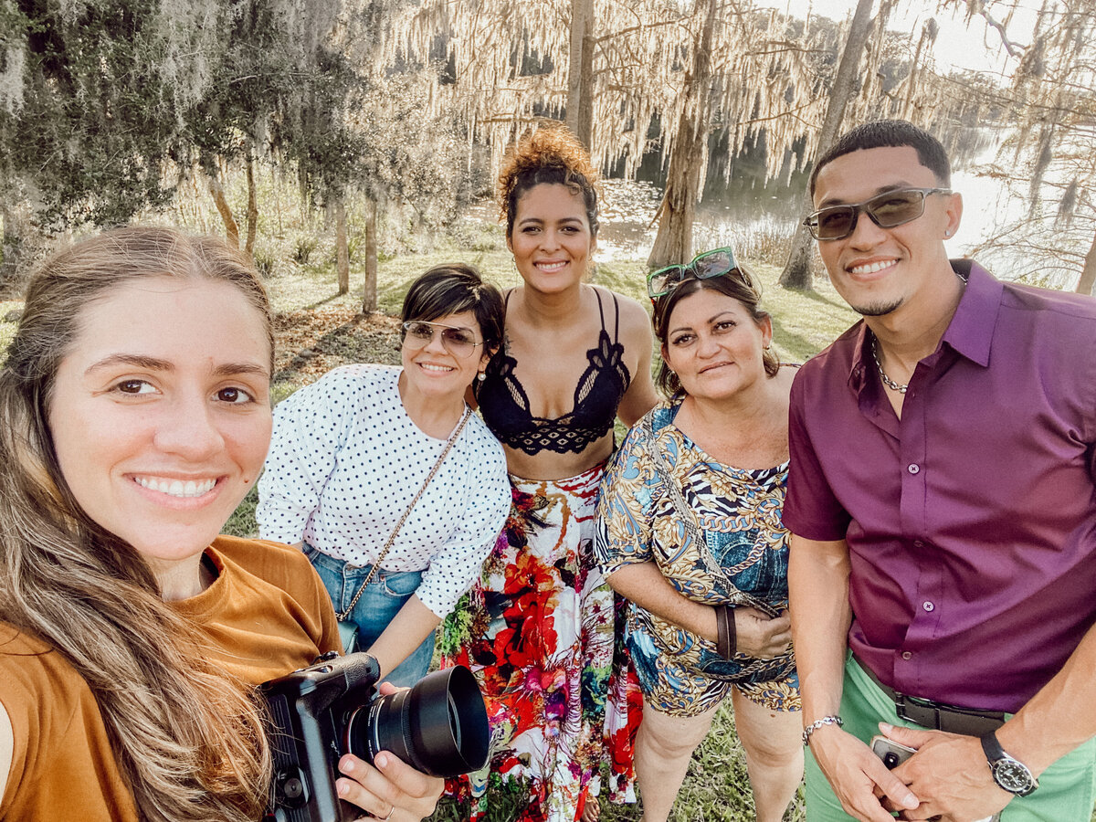 Smiling photographer with an engaged couple and family after an outdoor engagement session surrounded by Spanish moss trees.