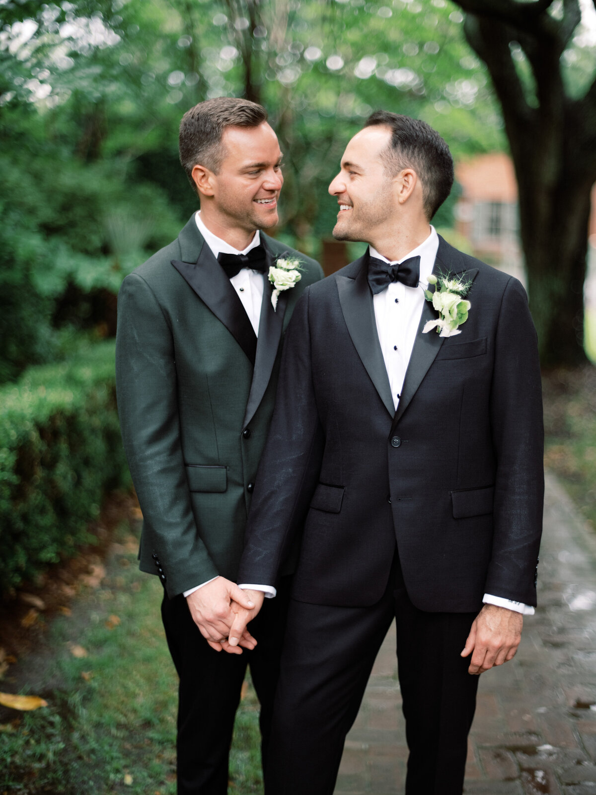 Two grooms stand close together holding hands, displaying their love and affection, surrounded by a picturesque outdoor setting that emphasizes their joyous union.