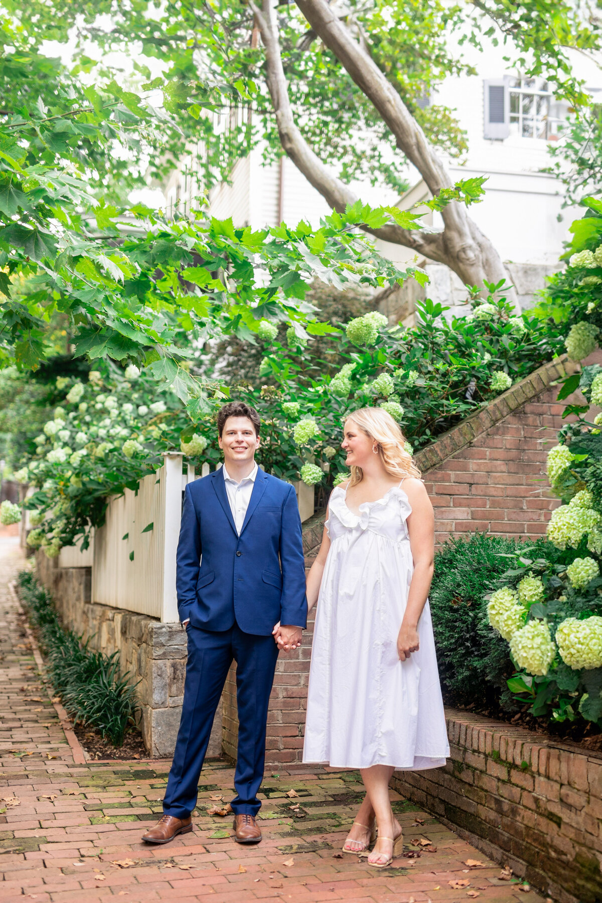 A couple stands hand in hand on a brick pathway surrounded by lush greenery and hydrangea flowers. The man is wearing a blue suit, and the woman is in a white summer dress, both smiling outdoors on a sunny day.