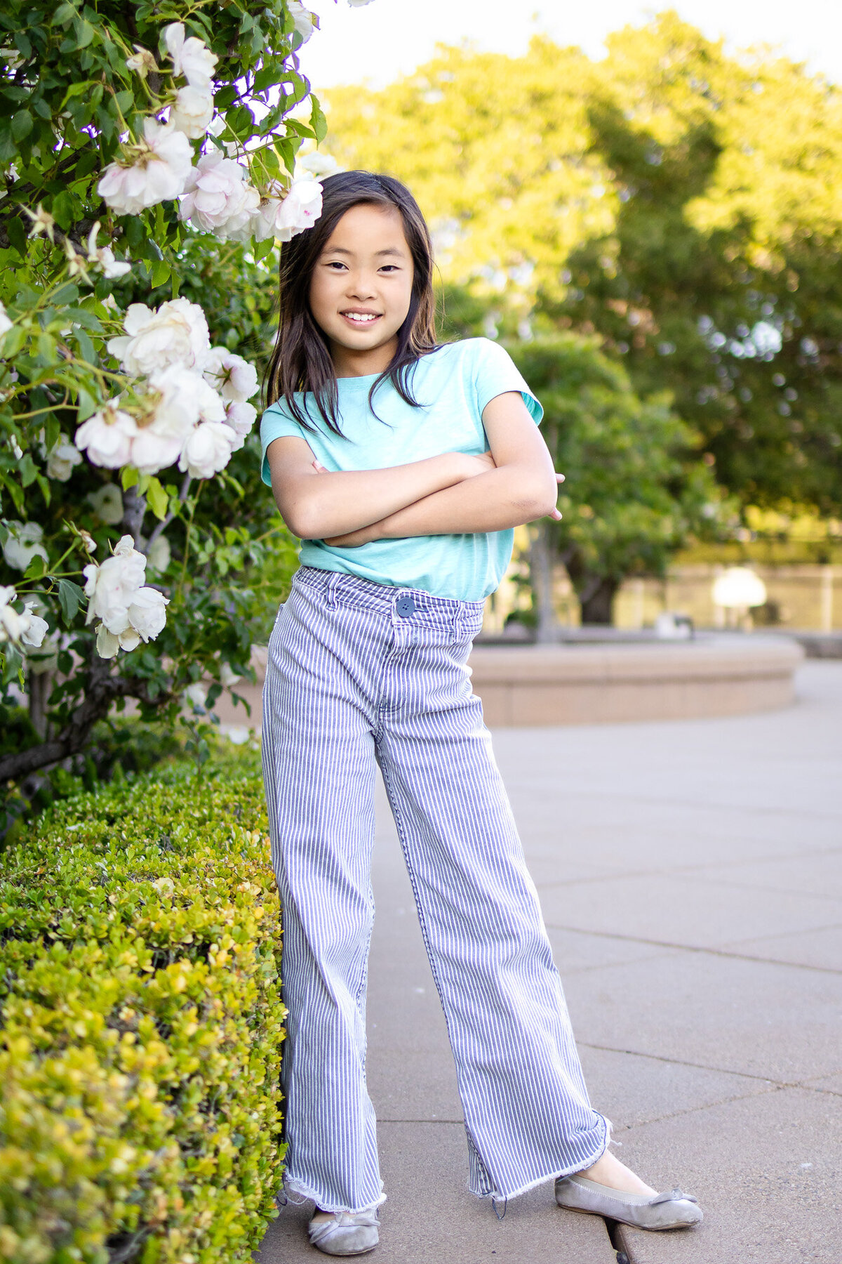 Confident young girl smiling with arms crossed in front of blooming white roses – Bay Area Family Portfolio - Ellobelle Photography