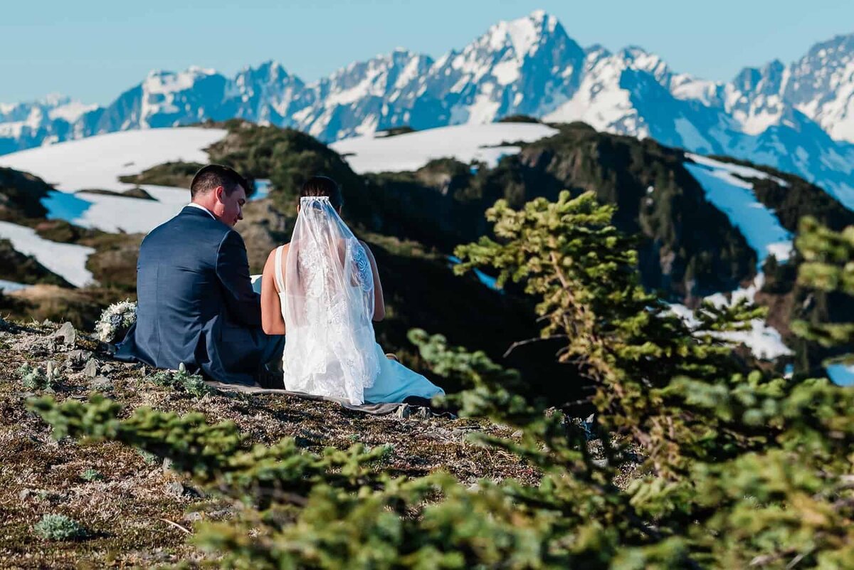 A bride and groom in their wedding finest sit atop a mountain summit of flat rocks covered in tundra grass.  behind them the view drops off to ultra blue water of Resurrection Bay in Seward, Alaska.  There are mountains jagged, snow covered and laced with green shooting up out of the bay, in a spot only accessible via the helicopter they took during their private heli and glamping elopement package.  The bride's long white brushes the tundra as they read letters from home.
