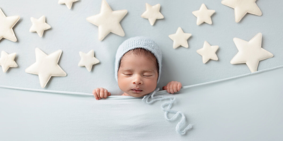Newborn baby boy wearing a light blue knit bonnet, tucked under a soft blue blanket with felt stars arranged above him.