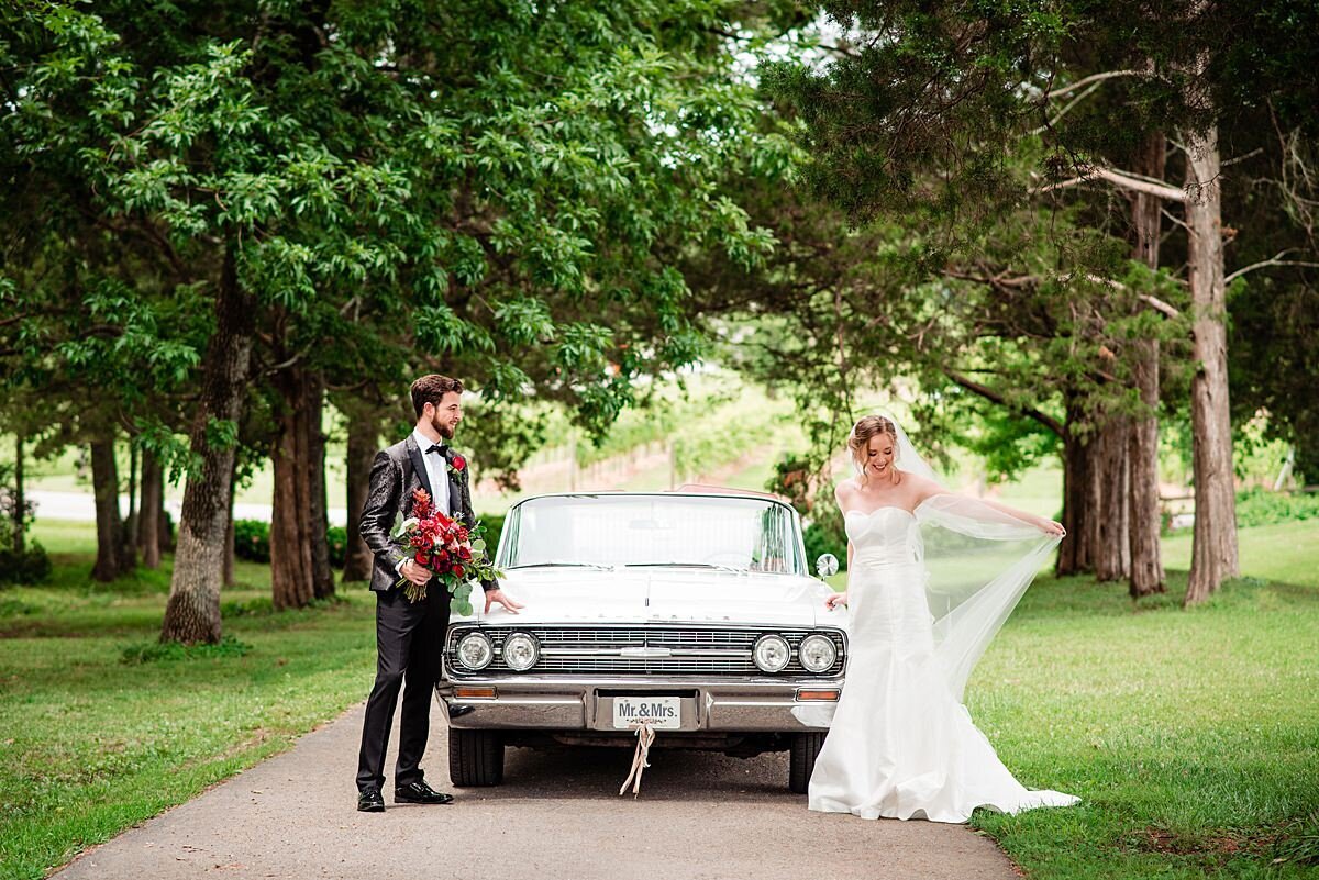 The bride, wearing a long silk wedding dress with a strapless lace bodice, sweetheart neckline and a long sheer veil and the groom wearing a black tuxedo with a black silk jacket embroidered with a paisley pattern and red rose boutonniere as they stand next to their vintage 1964 white Oldsmobile convertible. The groom is holding the bride's large red and blush bridal bouquet at their summer wedding at Arrington Vineyards.